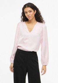 Blouse en satin rose clair avec un décolleté en V et des manches bouffantes à poignets élastiqués, associée à un pantalon noir taille haute.