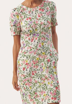 Femme portant une robe blanche longueur genoux avec des manches courtes et un motif floral coloré comprenant des fleurs roses, rouges, jaunes, violettes et vertes.