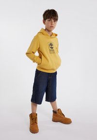 Sweat-shirt à capuche jaune avec logo, shorts en denim bleu et bottes de randonnée marron. La tenue présente des éléments de design décontractés et confortables.