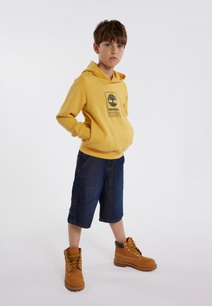 Sweat-shirt à capuche jaune avec logo, shorts en denim bleu et bottes de randonnée marron. La tenue présente des éléments de design décontractés et confortables.