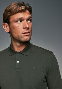 Polo de color verde oscuro con tejido texturizado, tapeta de tres botones y un pequeño logo bordado en el lado izquierdo del pecho.