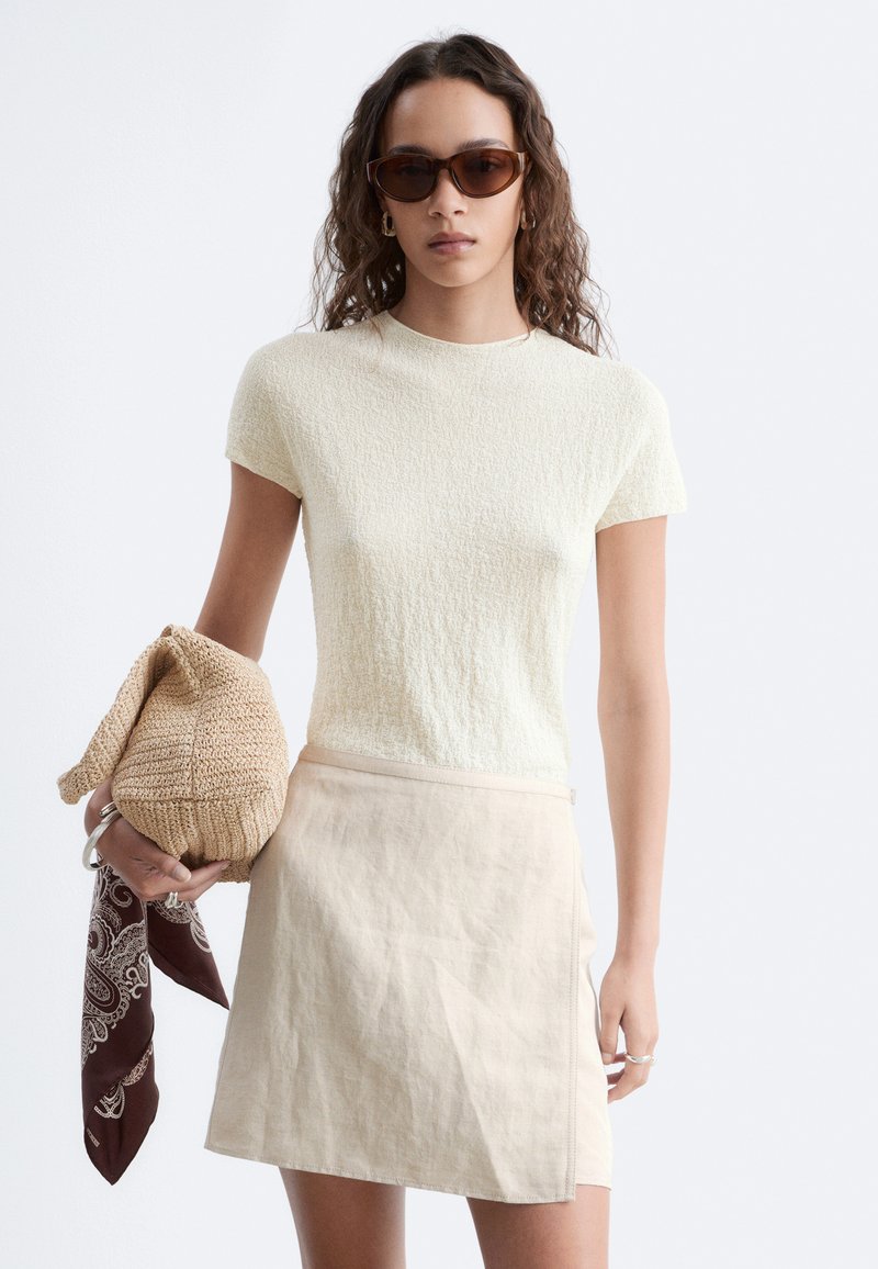 Femme aux cheveux bouclés portant des lunettes de soleil marron, un haut crème à manches courtes, une jupe beige, tenant un sac tricoté avec une écharpe marron.