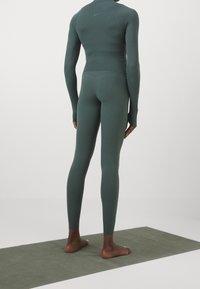 Długi rękaw sportowy top i legginsy w ciemnozielonym materiale, dopasowany krój, gładka faktura, minimalne logo, noszone na bosą stopę na macie do jogi.