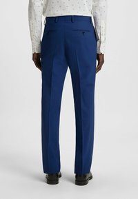 Blauwe op maat gemaakte broek met een gladde afwerking, voorzien van twee achterzakken met knopen en een plooi detail langs de benen. Zwarte schoenen zichtbaar.