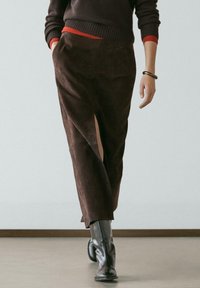 Person trägt einen braunen Wildlederrock mit vorderem Schlitz, schwarze Lederstiefel, einen dunklen Pullover über einem roten Hemd und ein Armband und geht drinnen.