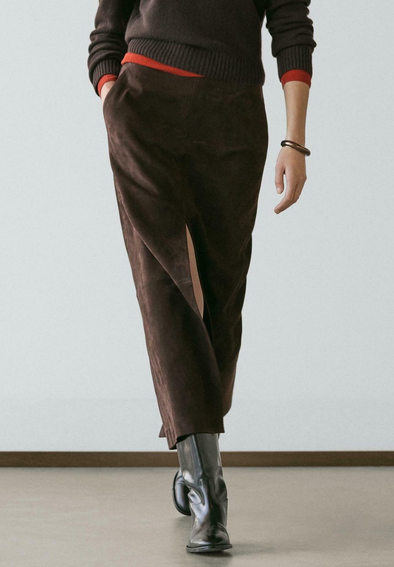 Person trägt einen braunen Wildlederrock mit vorderem Schlitz, schwarze Lederstiefel, einen dunklen Pullover über einem roten Hemd und ein Armband und geht drinnen.