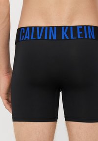 Boxeri negri din material elastic, cu un logo „CALVIN KLEIN” de culoare albastră pe talie. Material neted, design ajustat și lungime scurtă, deasupra coapsei.