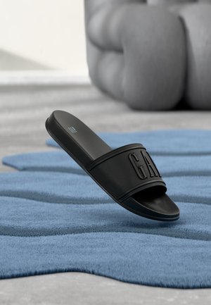 Sandale noire à glissière avec logo "GAP" en relief sur la sangle, posée sur un tapis bleu texturé, avec un grand objet gris flou en arrière-plan.