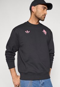 Felpa nera con scollo rotondo, maniche lunghe e polsini a coste. Presenta dettagli logo rosa sul petto e sulla parte superiore sinistra.