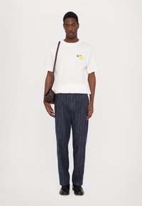 Witte katoenen t-shirt met een gele graphic, gecombineerd met donkerblauwe gestreepte broek. Een kleine bruine schoudertas maakt de look compleet.