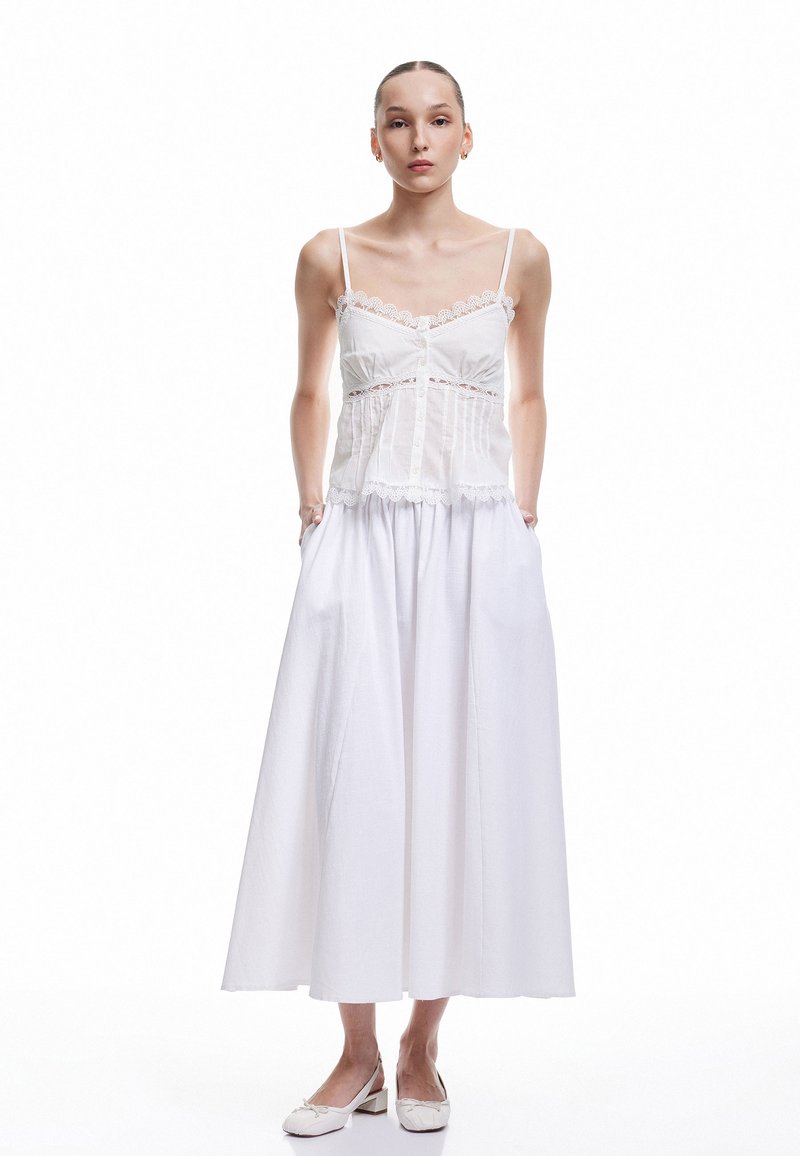 Camiseta blanca sin mangas con detalles de encaje y botones, combinada con una fluida falda larga blanca, hecha de tela ligera. Acentuada con zapatos blancos.