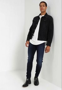 Črna jeans jakna preko bele srajce, v kombinaciji z temno modrimi oprijetimi kavbojkami in črnimi usnjenimi gležnjarji. Minimalistična zasnova in kroj.