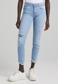 Jeans de mezclilla azul claro con detalles desgastados y dobladillo deshilachado. Combinados con zapatillas blancas, mostrando un diseño casual y ajustado.