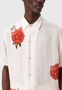 Persona de piel oscura con una camisa blanca de manga corta abotonada, con grandes estampados de rosas rojas en el pecho y el dobladillo inferior.