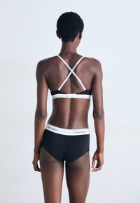 Svart bralette med X-bakstropper og justerbar spenne, kombinert med svarte hipster-truser som har en hvit Calvin Klein-logo midjebånd.