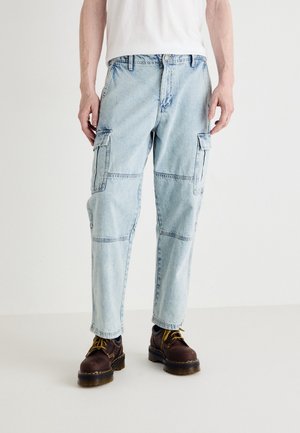 Man draagt lichtblauwe denim cargobroek met grote zijzakken, donkerbruine leren laarzen met gele veters en een wit shirt.