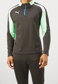 Långärmad träningsjacka i svart tyg med gröna detaljer, med halvdragkedja och blå dragkedja, samt en vit logotyp på bröstet.