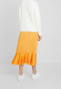 Gonna in satin arancione con orlo svasato, abbinata a un maglione bianco in maglia e sneakers bianche. Il tessuto ha una texture liscia.