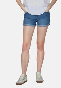 Shorts de mezclilla en azul medio con dobladillos enrollados, con bolsillos y un diseño ajustado, combinados con zapatillas blancas y una blusa blanca suelta.