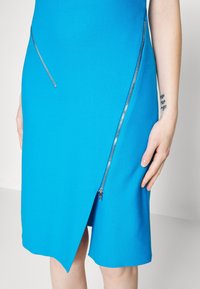 Tyrkysové šaty s přiléhavým střihem, zdobené stříbrnými zipy a asymetrickým lemem. Hladká textura s výraznou strukturou.