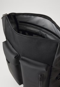 Juoda kuprinė su užtrauktuku viršuje, tekstūruotu paviršiumi, priekinėmis kišenėmis ir vidinėmis akių skyriais. Lygus audinys su tvirtomis detalėmis.