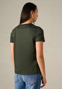 T-shirt vert foncé à manches courtes en coton texturé, avec un col rond et une coupe standard, associé à un jean en denim bleu clair.