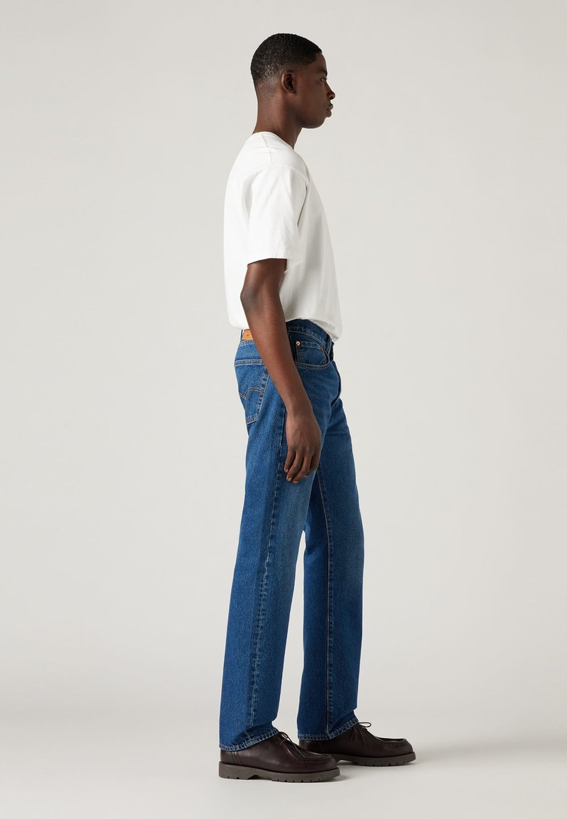 Blaue Jeans im geraden Schnitt, kombiniert mit einem weißen Rundhals-T-Shirt. Schwarze Schnürschuhe ergänzen das Outfit.