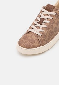 Baskets marron à motif avec lacets blancs, semelle blanche et étiquette de marque en cuir près des œillets supérieurs sur fond blanc.
