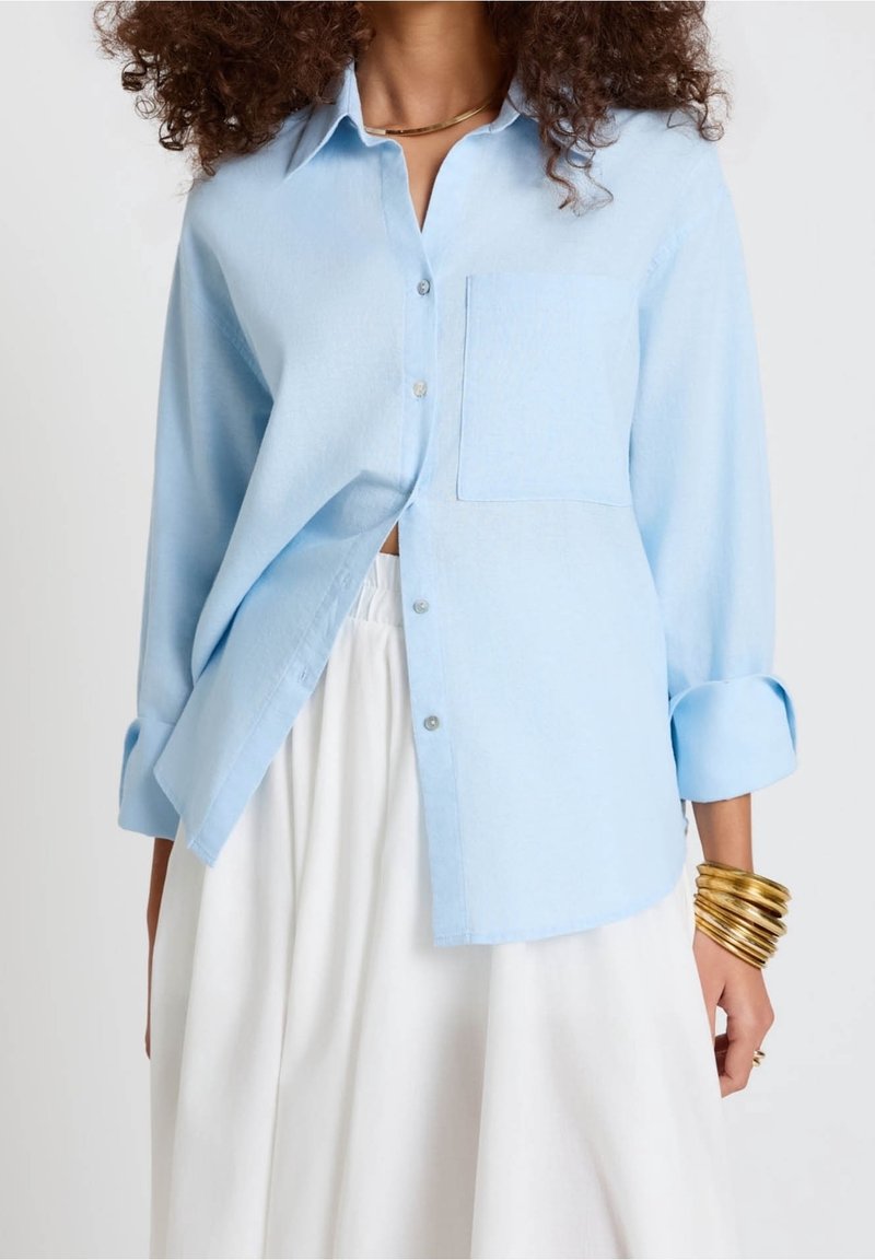 Femme portant une chemise bleu clair à boutons avec des manches retroussées, une jupe blanche taille haute, des bracelets en or et un collier en or.