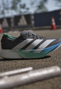 Scarpa da corsa grigia con strisce bianche e suola blu-verde posizionata sull'asfalto vicino a coni stradali e barriere sfocate.