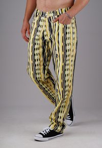Man wearing yellow, black, and white patterned pants, black sneakers with white laces, and a silver bracelet with one hand in pocket.
