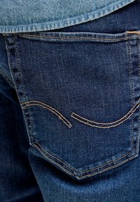 Zbliżenie na tylną kieszeń niebieskich dżinsów z charakterystycznym zakrzywionym przeszyciem i widocznymi szwami.