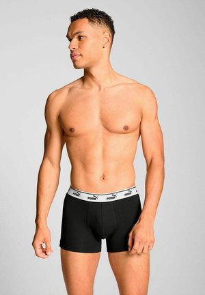 Muskelbepackter, oberkörperfreier Mann steht, schaut nach links, trägt schwarze Puma-Boxershorts mit weißem markierten Bund vor einem schlichten grauen Hintergrund.