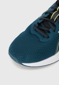 Chaussure de sport teal avec tige en mesh, surface texturée et lacets noirs. Présente des accents jaunes et une semelle blanche avec une légère bordure bleue.