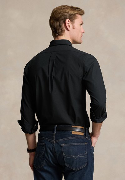 Man zijwaarts kijkend met een zwart shirt met lange mouwen en donkerblauwe spijkerbroek, handen bij de zakken, tegen een effen beige achtergrond.