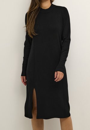 Woman wearing a long-sleeve black midi dress with a crew neck and front slit, standing against a plain light background.
