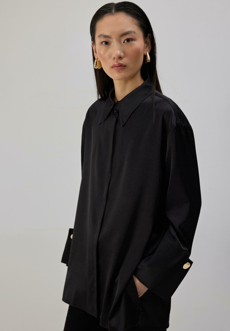 Γυναίκα με μακριά σκούρα μαλλιά που φοράει υπερμεγέθη μαύρο πουκάμισο με κουμπιά και χρυσά γεωμετρικά σκουλαρίκια, στέκεται μπροστά σε ένα απλό ανοιχτό γκρι φόντο.