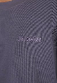 Pull violet à côtes avec un tissu texturé, portant "Dropsize" brodé dans une couleur mauve claire contrastante sur la poitrine.