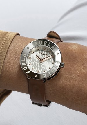 Montre argentée avec un cadran rond, présentant un cadran à motif clair et un bracelet en cuir marron. "FURLA" gravé autour de la lunette.