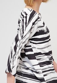 Blouse rayée noir et blanc avec des manches trois-quarts, offrant une coupe ample et des motifs de coups de pinceau abstraits sur un tissu léger.