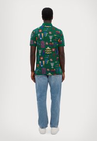 Zielona koszulka polo z powtarzającym się wzorem pamiątek sportowych, trofeów i koszulek; połączona z jasnoniebieskimi dżinsami i białymi butami.