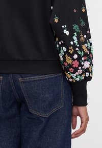 Sweat-shirt noir avec broderie florale colorée sur les manches ; présente des poignets froncés, un ourlet arrondi, et un jean en denim visible en dessous.