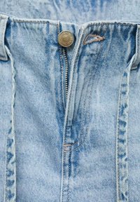 Svijetloplava denim tkanina koja čini pojas traperica, s metalnim dugmetom i zatvaračem, vidljivim detaljima šivanja.