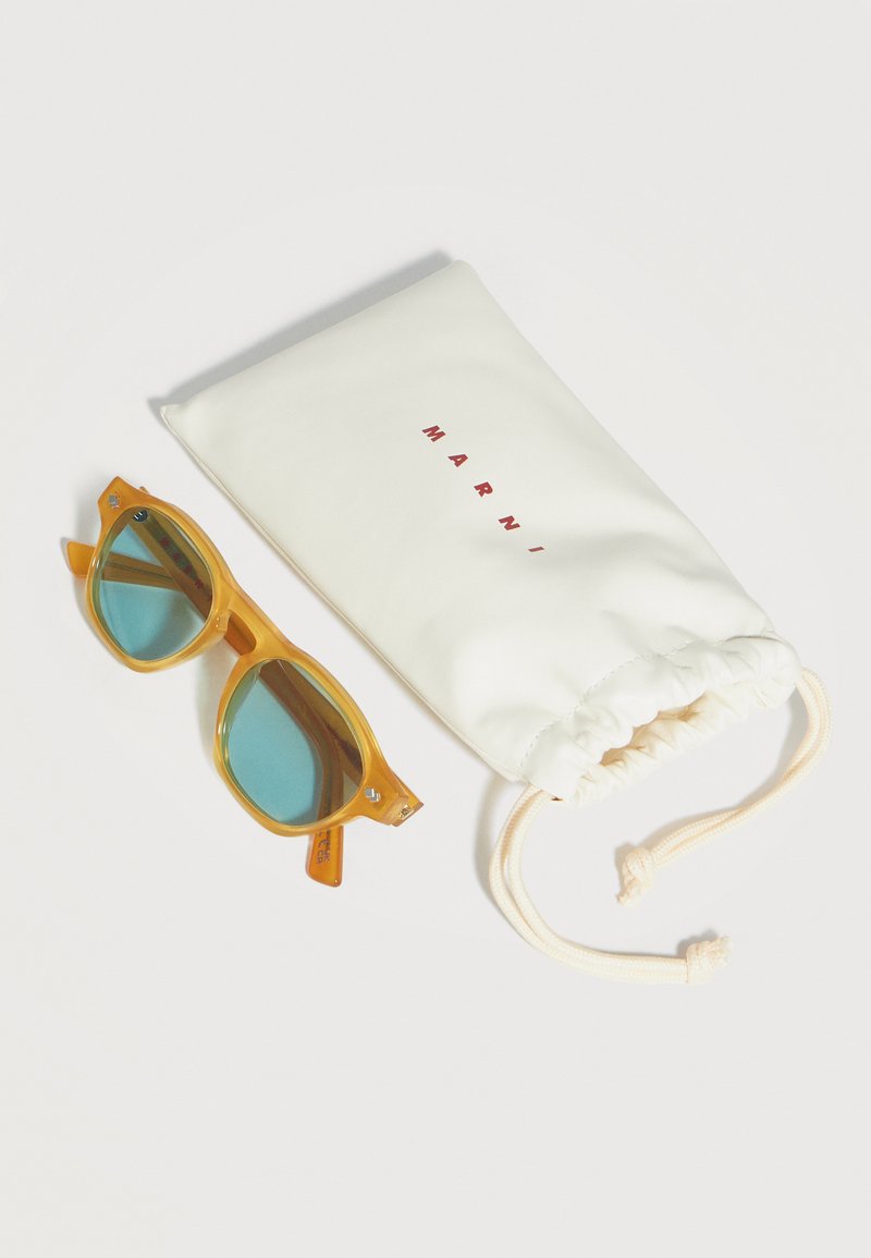 Occhiali da sole con montatura gialla e lenti blu accanto a una bustina bianca con coulisse etichettata "MARNI" su una superficie bianca liscia.