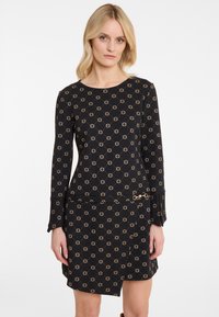 Frau trägt ein schwarzes Langarmkleid mit beigen geometrischen Mustern und einem asymmetrischen Saum vor einem schlichten weißen Hintergrund.