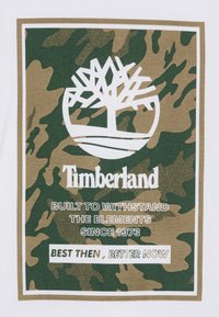 Valge T-särk, millel on roheline ja pruun kamuflaažiprint. Keskosa logo valge puu ja "Timberland" tekstiga, millele on lisatud sloganid.