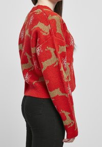 Roter Strickpullover mit einem Hirschmuster in Gold und Weiß, mit rundem Halsausschnitt und gerippten Bündchen, kombiniert mit schwarzen Hosen.