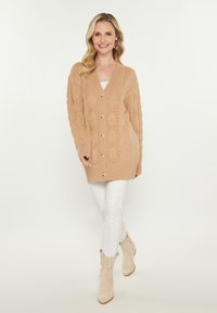 Beige Strickjacke mit V-Ausschnitt, die mit Rautenmuster und Knopfakzenten versehen ist. Knopfverschluss mit gerippten Bündchen und Saum; kombiniert mit weißen Hosen.