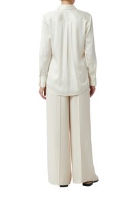 Cream-colored silk blouse with a back pleat, long sleeves, and a rounded hem, paired with wide-leg matching trousers.