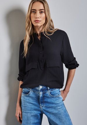 Camicia nera con bottoni, due tasche sul petto, tessuto texturizzato e maniche arrotolate, abbinata a jeans di denim blu.